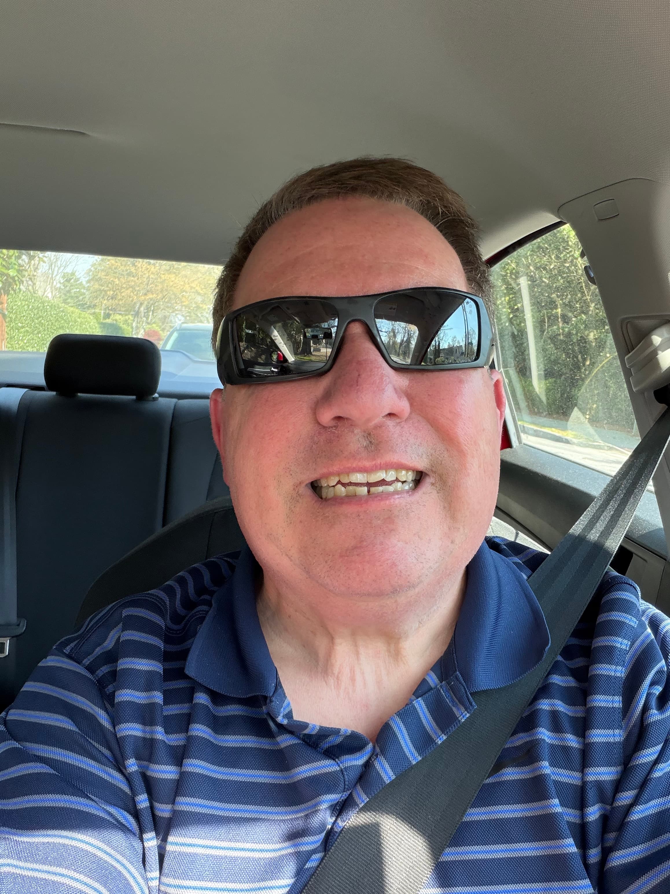 Smiling man wearing sunglasses and a blue striped polo shirt inside a car with seatbelt.